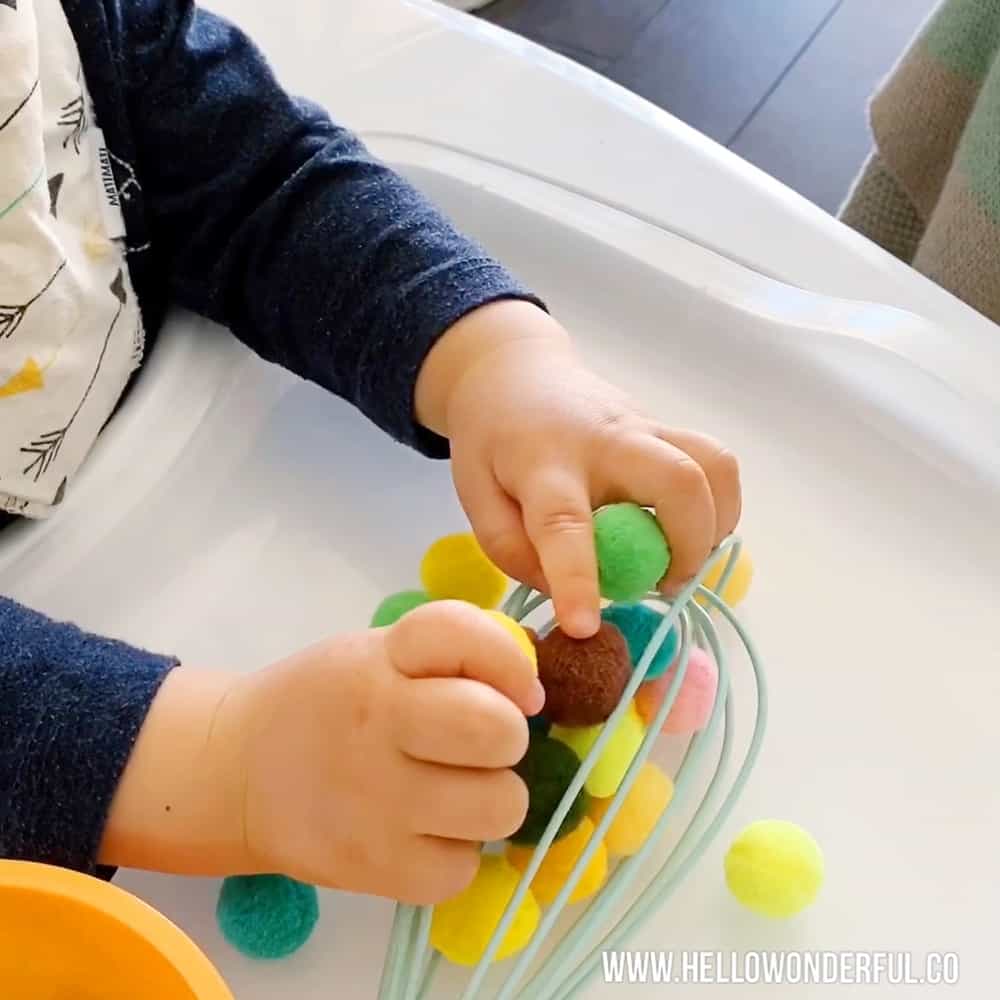 Baby playing with pom poms and kitchen whisk to practice fine motor and sensory skills. 