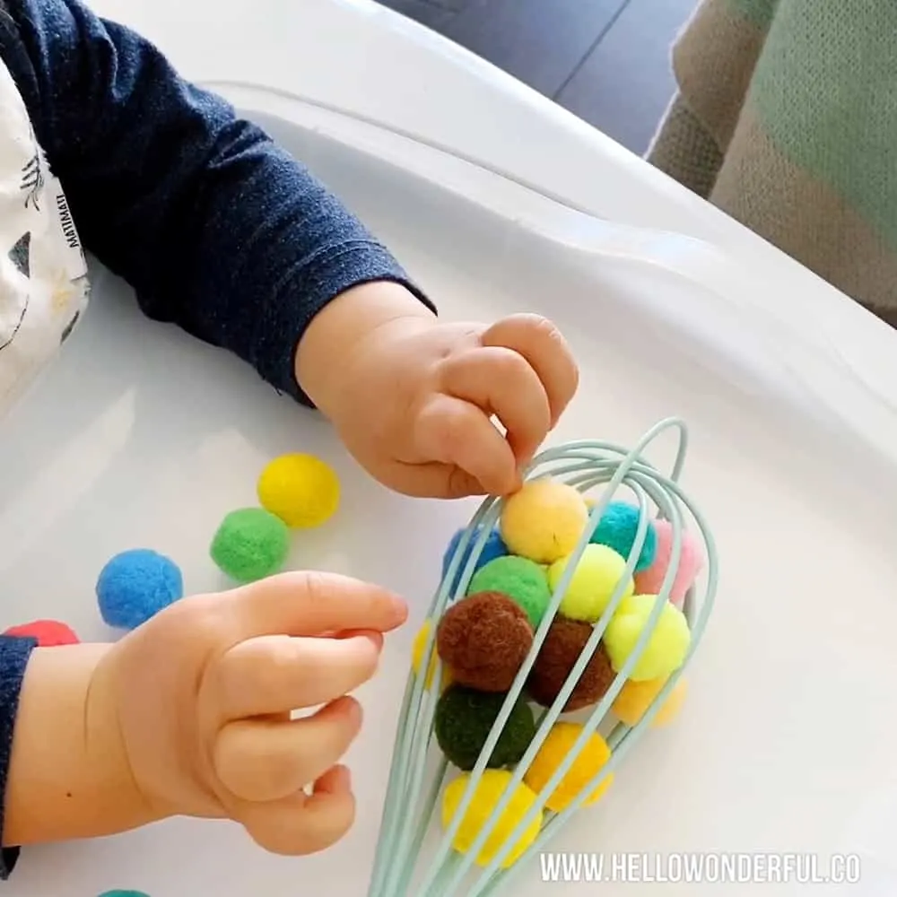 Baby playing with pom poms and kitchen whisk to practice fine motor and sensory skills. 