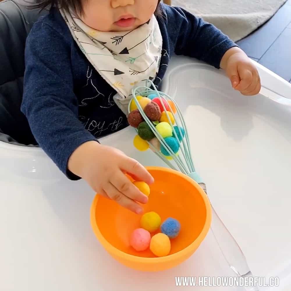 Baby playing with pom poms and kitchen whisk to practice fine motor and sensory skills. 