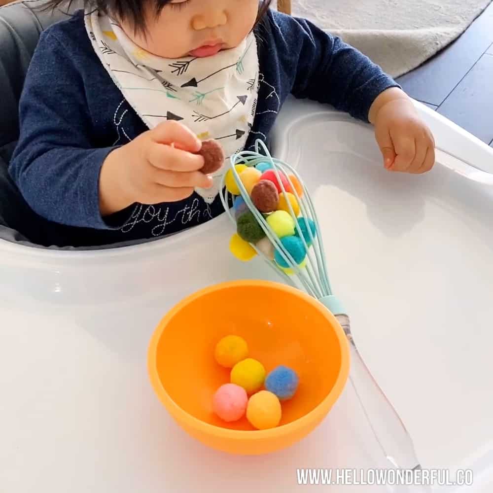 Baby playing with pom poms and kitchen whisk to practice fine motor and sensory skills. 