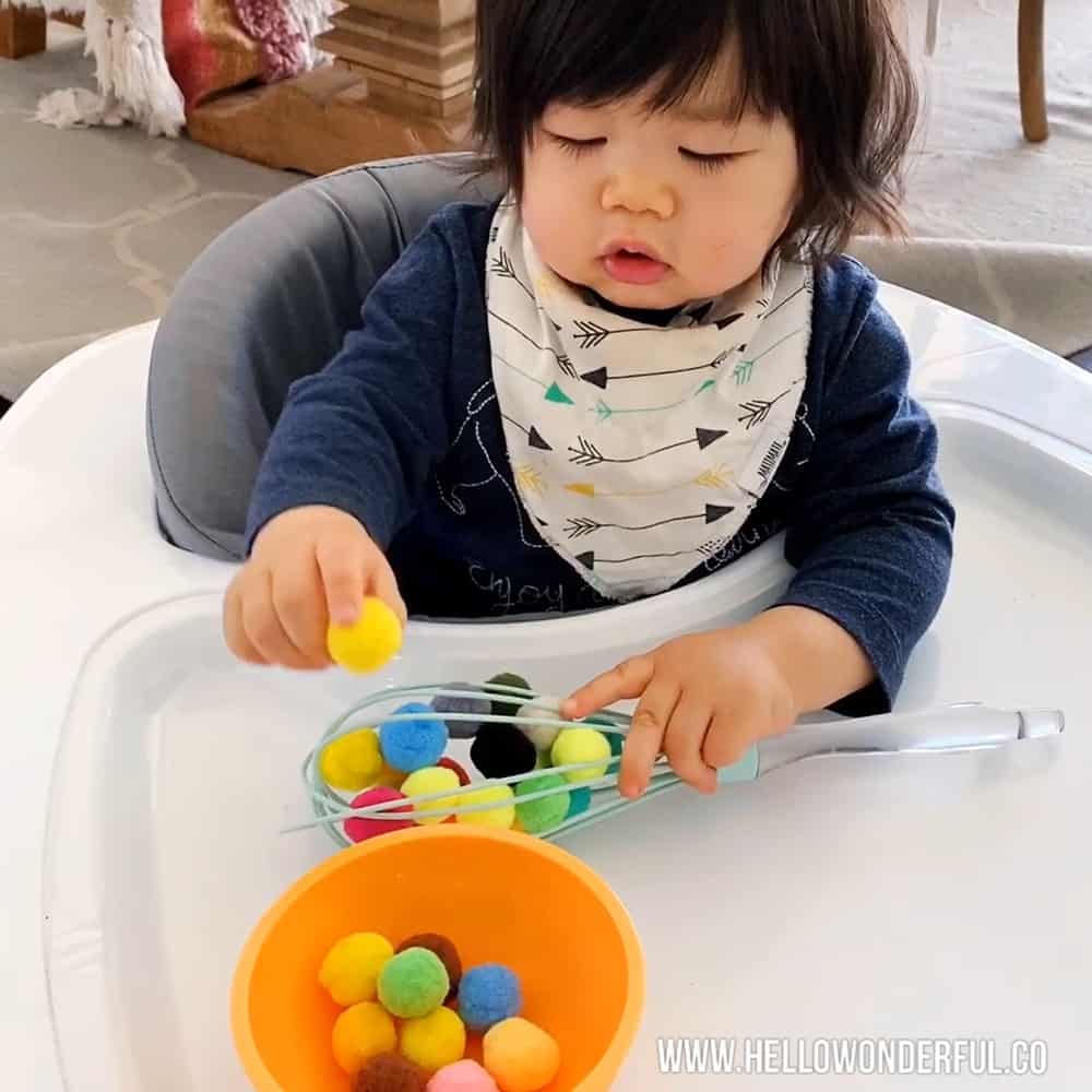 Baby playing with pom poms and kitchen whisk to practice fine motor and sensory skills. 