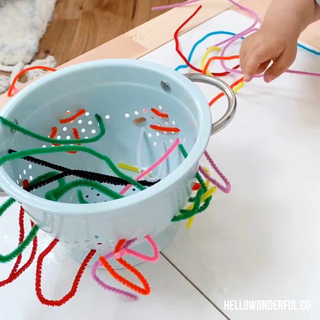 toddler grabbing pipe cleaners in colander to practice fine motor skills 