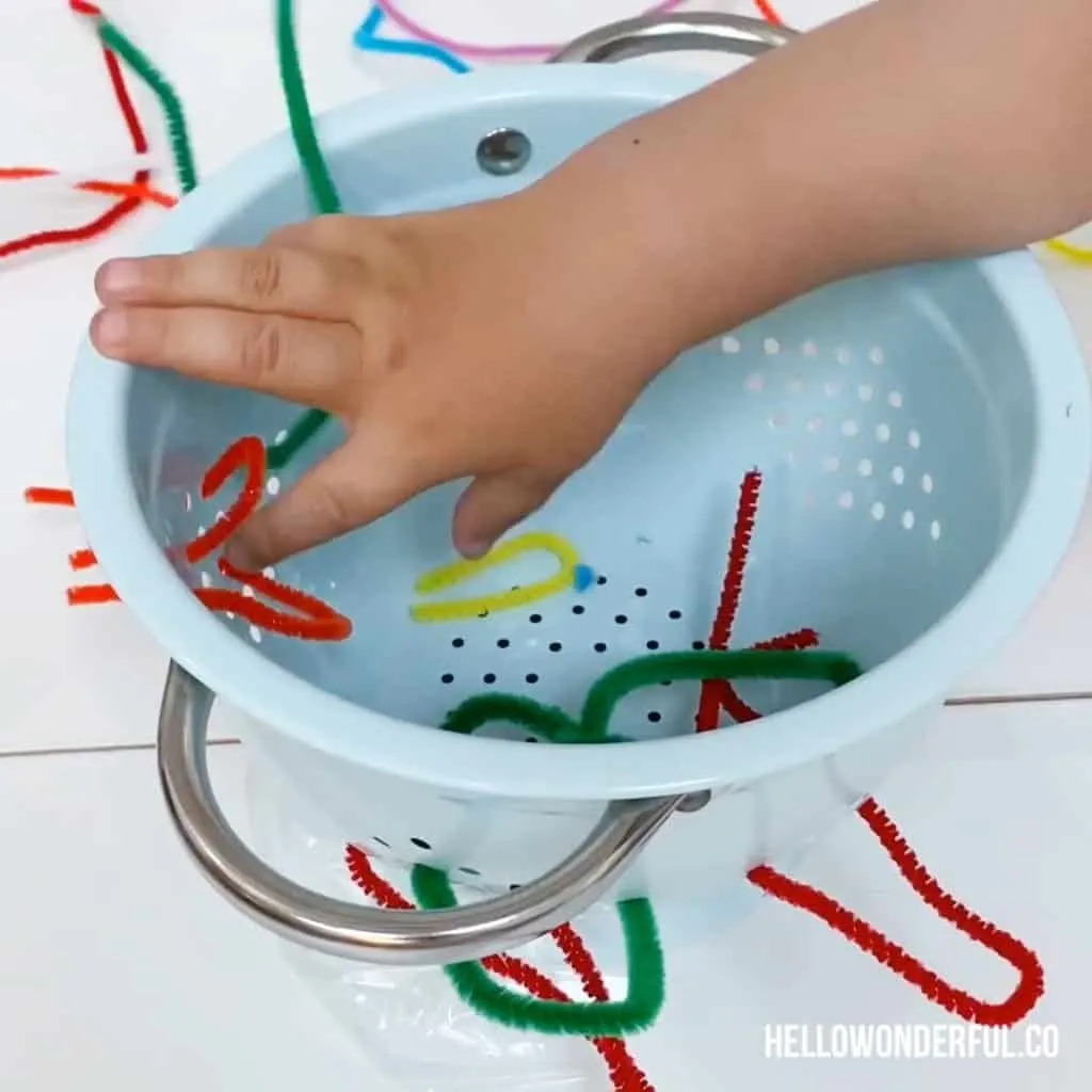 toddler grabbing pipe cleaners in colander for fine motor skills 